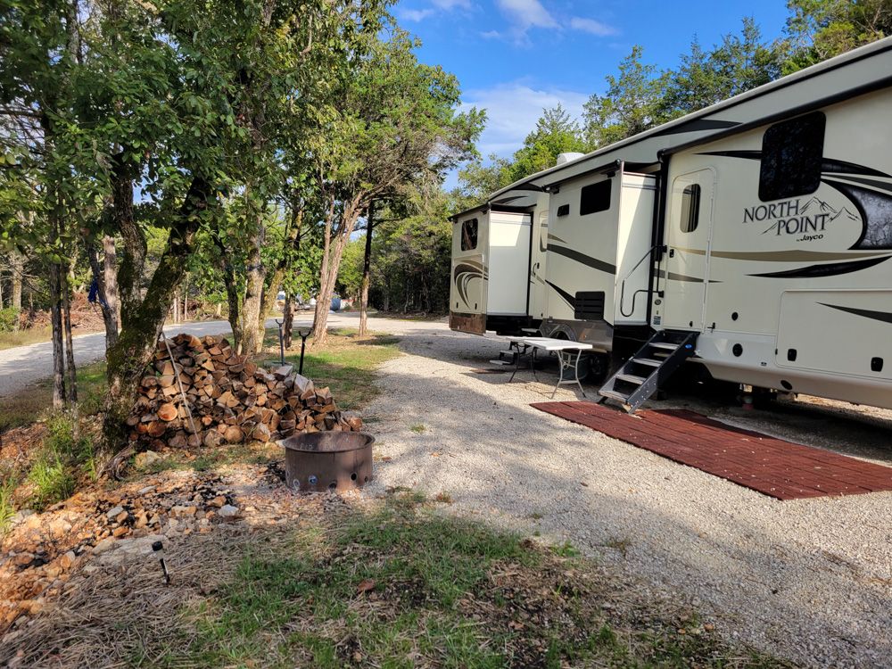 A North Point fifth-wheel RV on a level gravel site beside a stacked woodpile and a fire ring.