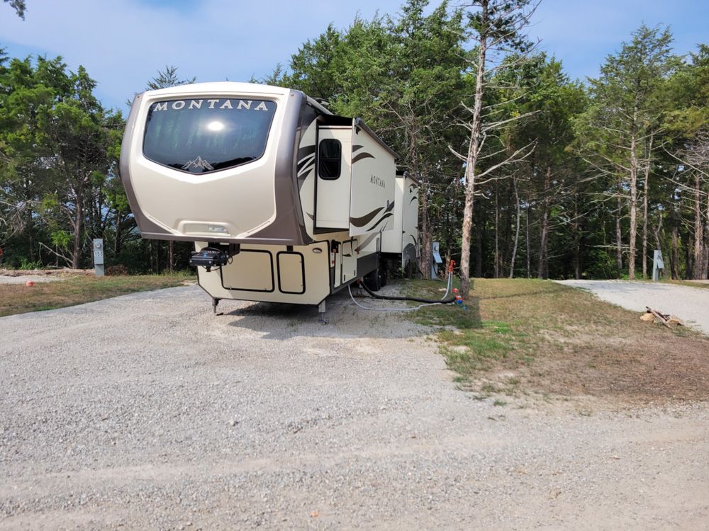 A Montana fifth-wheel RV set up on a spacious gravel site bordered by cedar trees.