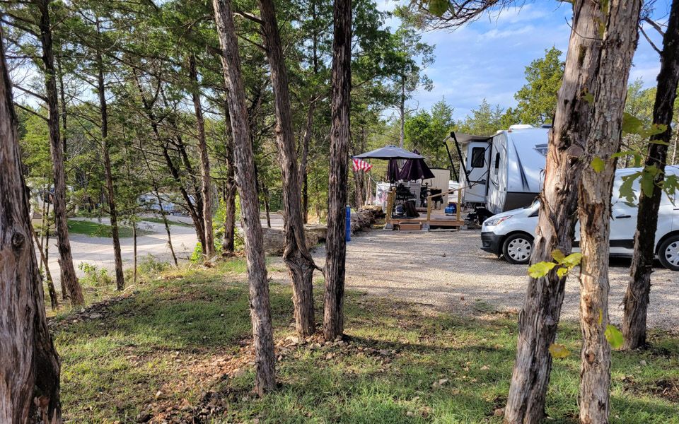 A shaded RV site tucked among cedar trees, with a travel trailer and a covered patio area.