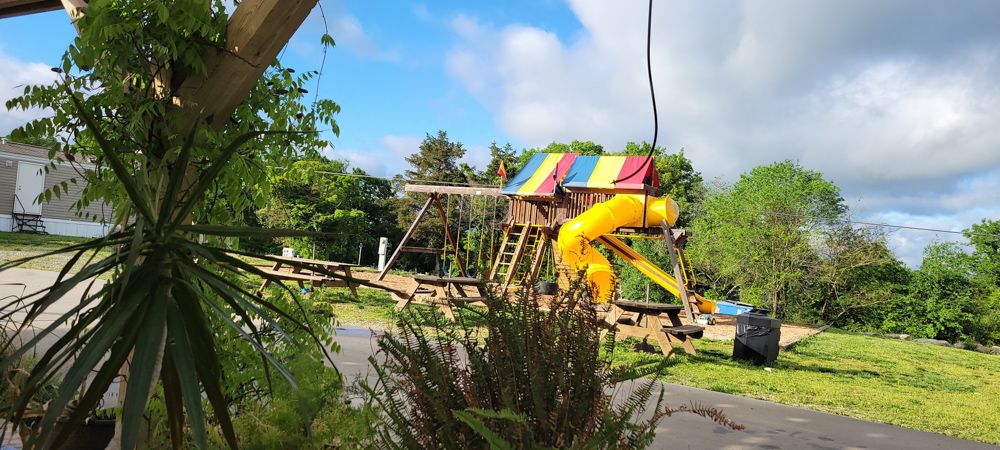 A children's playground with a yellow tube slide, swing set, and picnic tables on a sunny day.