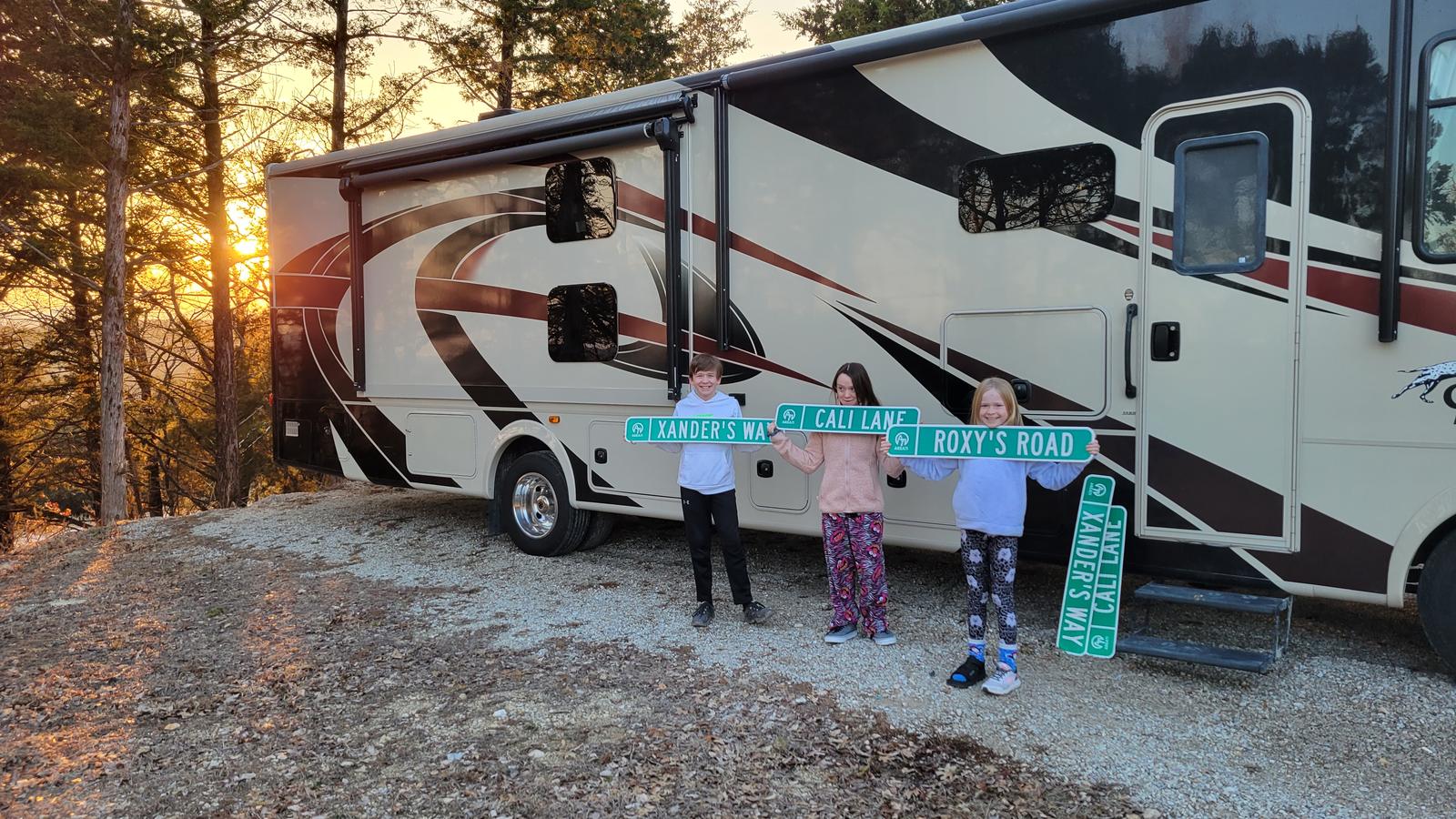 Three children standing in front of a motorhome at sunset, holding up green street
               signs reading Xander&rsquo;s Way, Cali Lane and Roxy&rsquo;s Road.